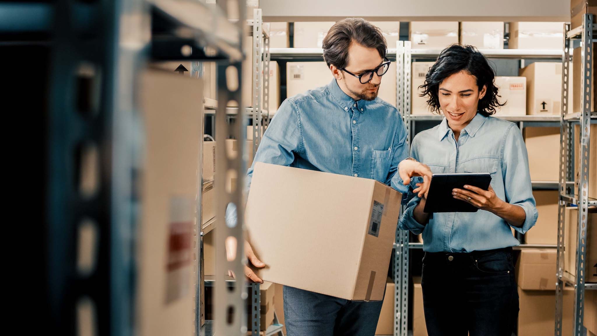 Female Inventory Manager Shows Digital Tablet Information to a Worker Holding Cardboard Box, They Talk and Do Work. In the Background Stock of Parcels with Products Ready for Shipment.