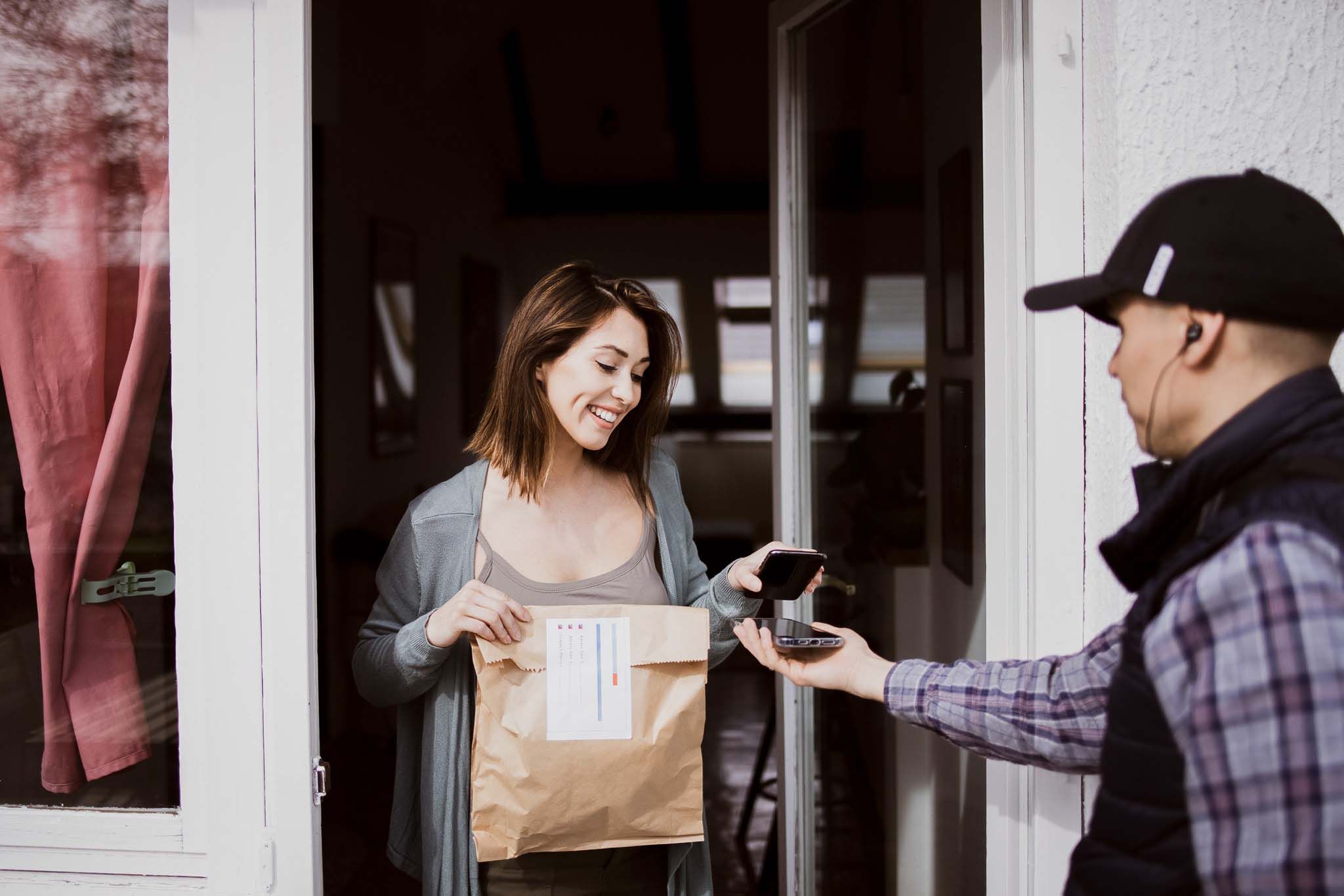 Young woman picking her order on doorstep from delivery guy and making contactless payment with phone
