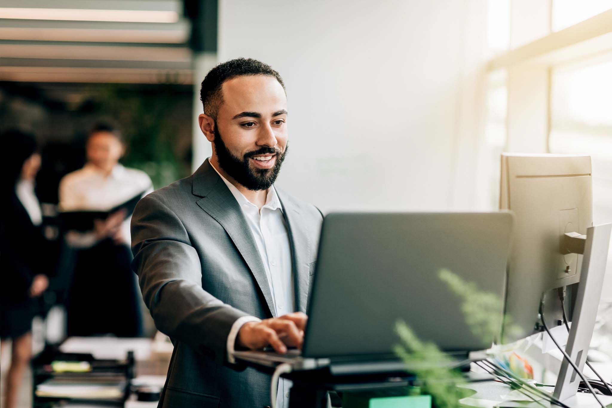 Multiracial Portuguese Jamaican mid adult businessman with beard standing smiling at desk with laptop reviewing data in bright business office wearing suit