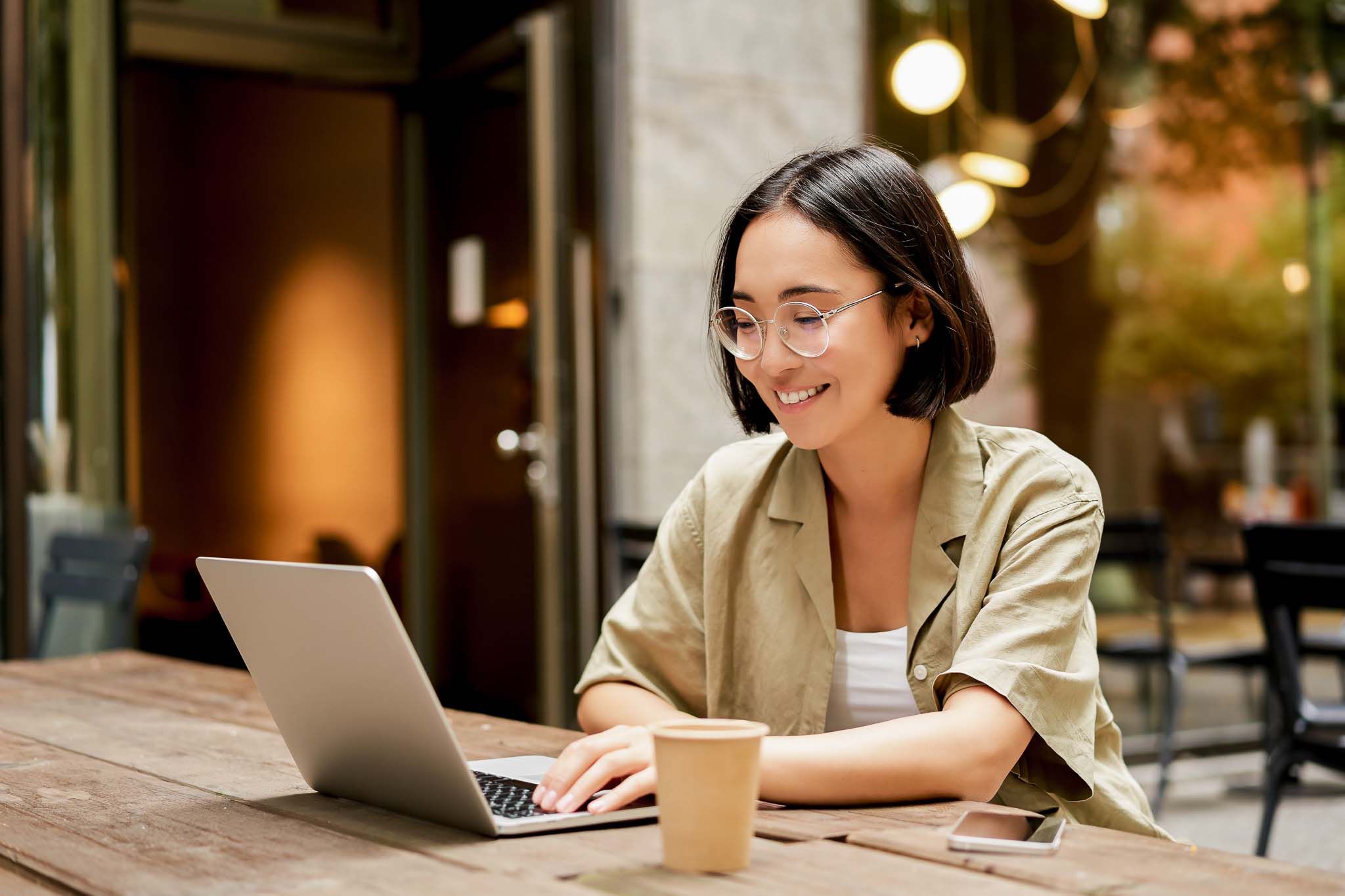 Young woman working in a cafe, using laptop and drinking coffee. Asian girl student with computer studying remotely, sitting on bench near shop